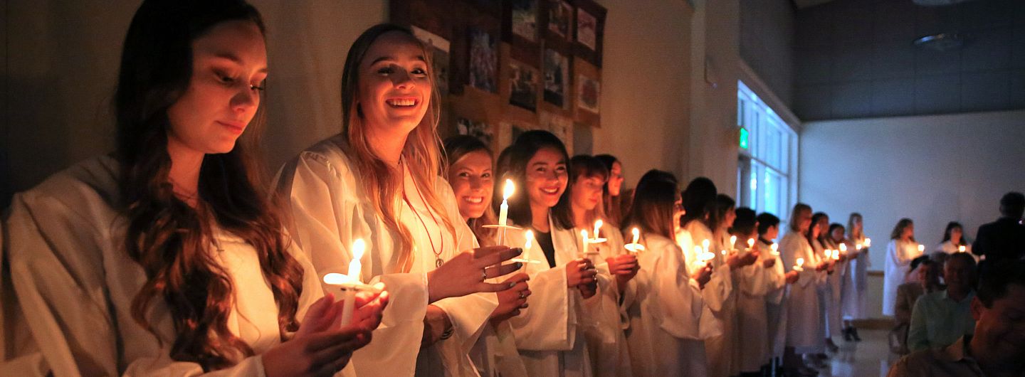A long line of students in grad gowns hold candles in the dark at Baccalaureate Mass on May 24, 2018.