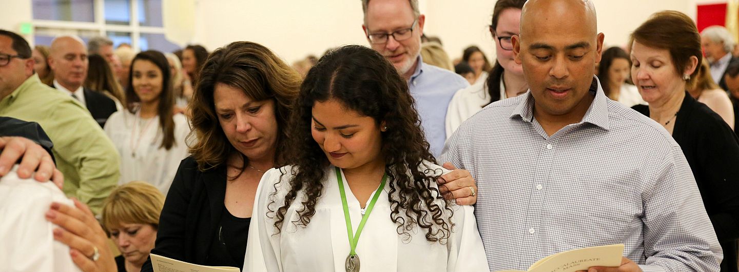 A student is flanked by a parent on either side, reading a prayer during Baccalaureate Mass on May 24, 2018.