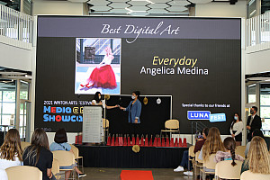 Students hands award to another student for her artwork on screen-- a woman in a red dress seated outside a coffee shop.
