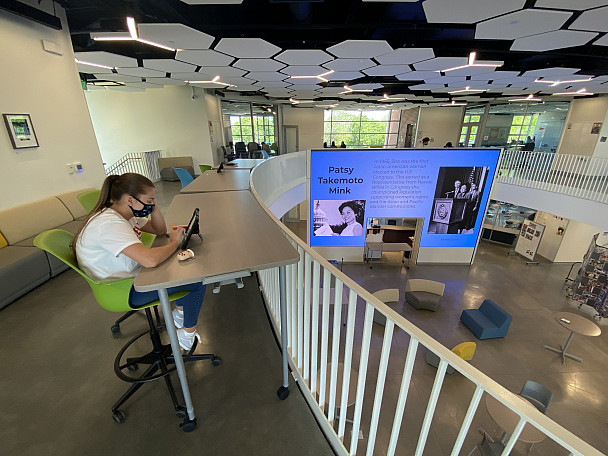 A student works at a table in the 2nd story balcony of the Innovation Center while the Media Wall...